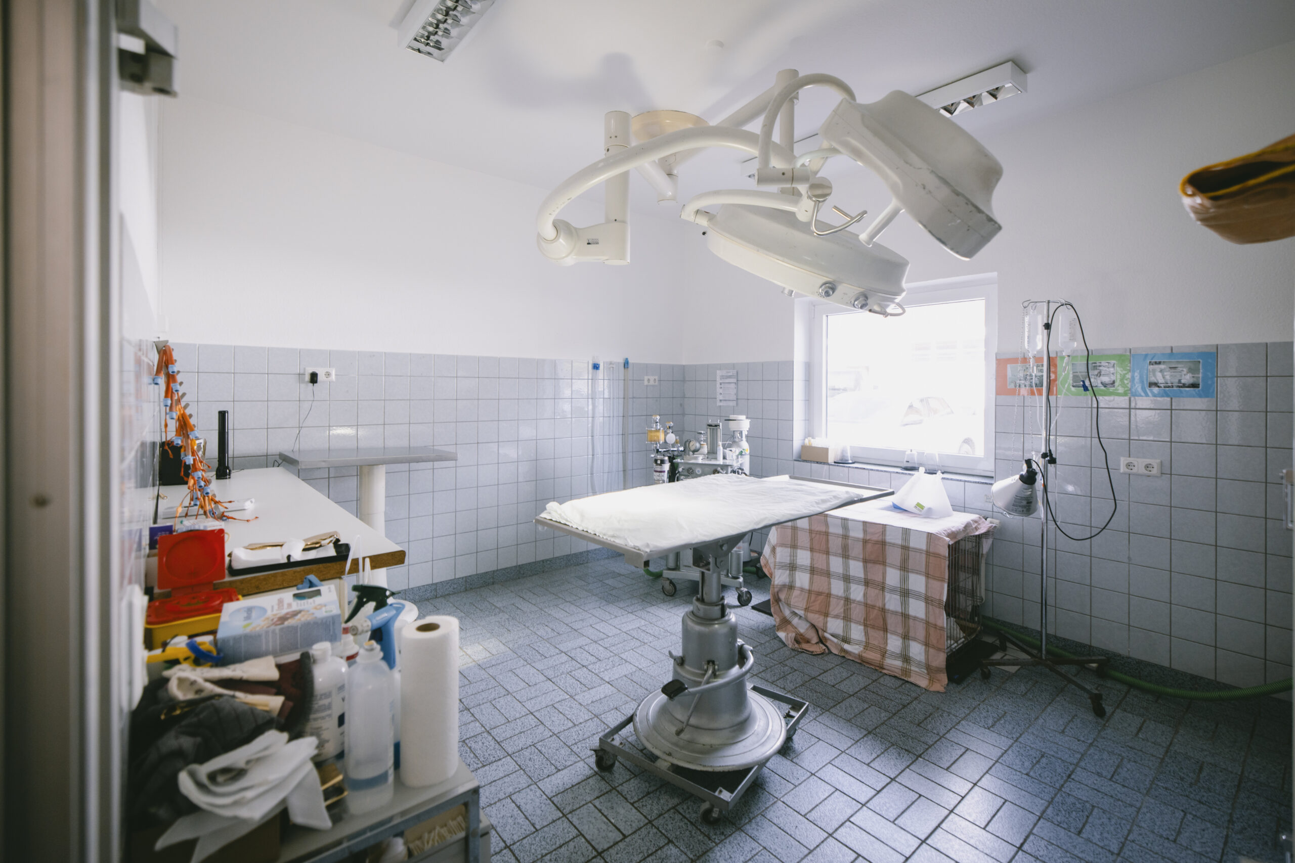 Operationsraum mit höhenverstellbarem Edelstahl-OP-Tisch, großer Deckenleuchte, Narkosegerät am Fenster, Infusionsständer und gefliesten Wänden.