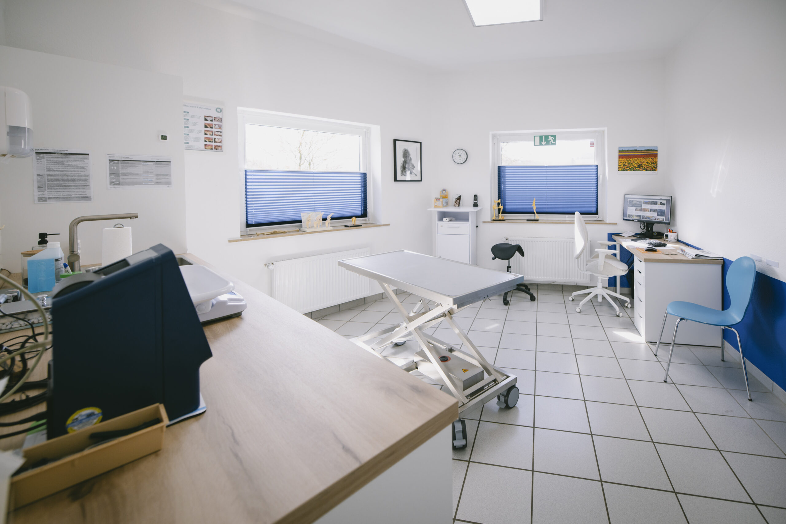 Behandlungszimmer mit Untersuchungstisch, Schreibtisch und blauen Jalousien an den Fenstern.